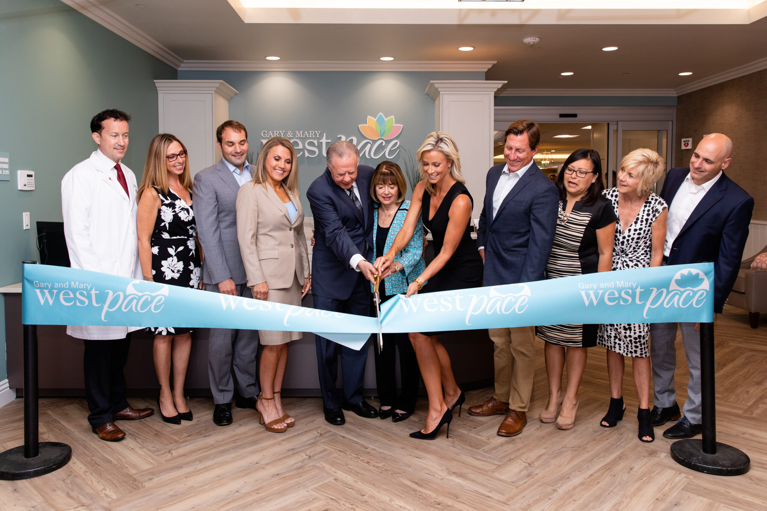 West Health & West PACE leadership teams, and San Marcos Mayor Rebecca Jones, posed next to Gary and Mary West, with large scissors, for the West PACE Grand Opening Ribbon Cutting Ceremony in lobby of West PACE day center.