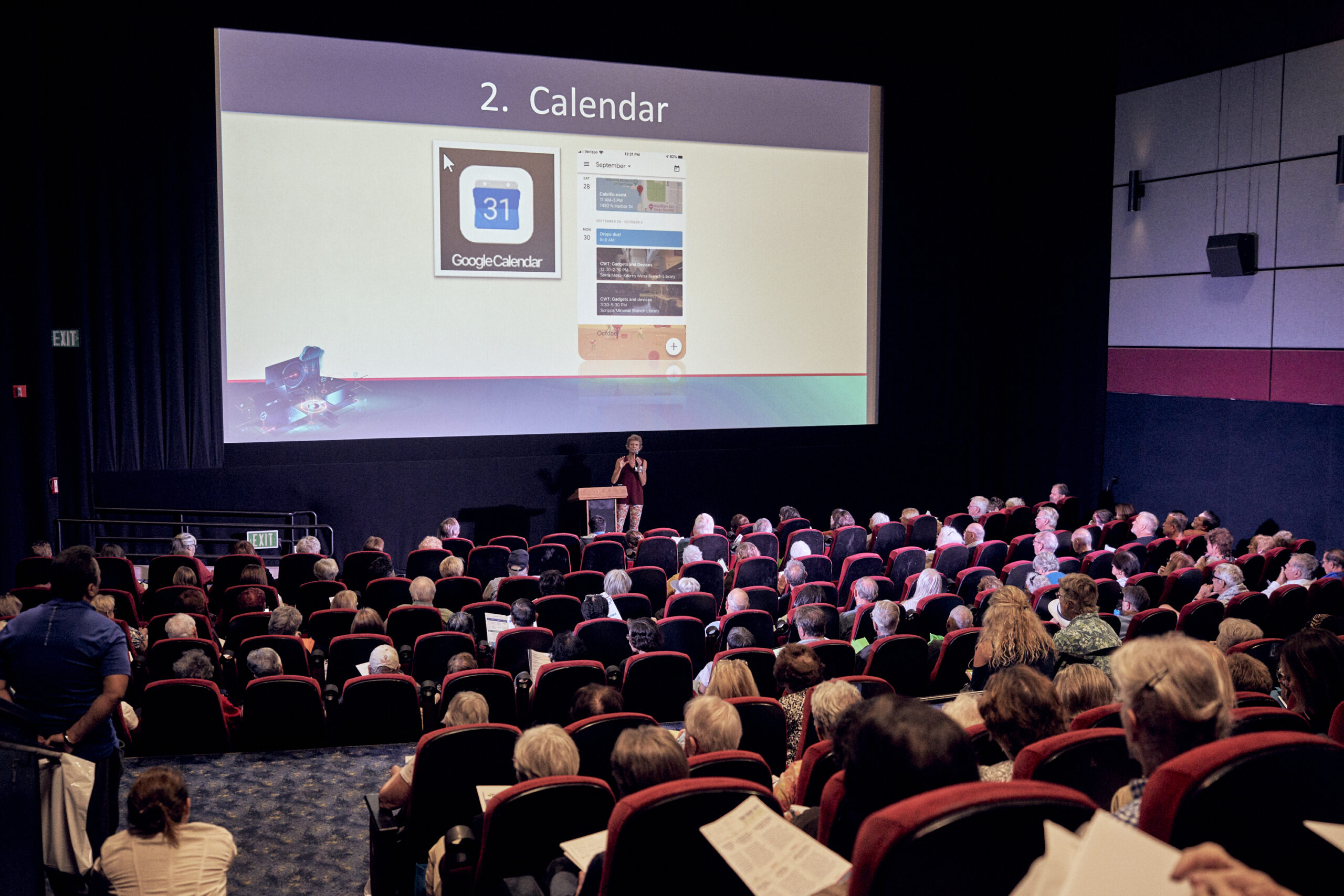 Cinmea theater full of seniors watching speaker present with projection screen showing Google Calendar on a mobile device.