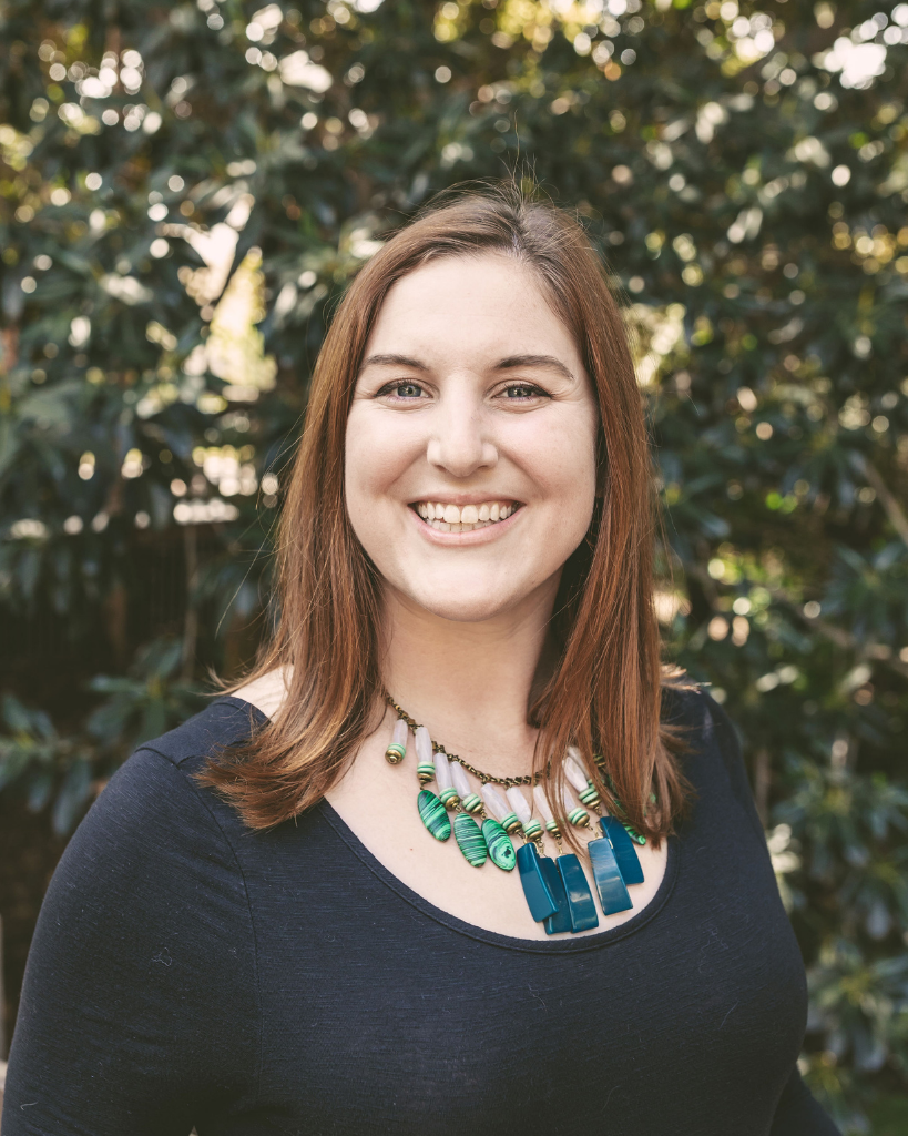 Scatena Daniels PR Consultant Sara Flocken photo in front of foliage wearing black top and decorative necklace with green and blue stones.