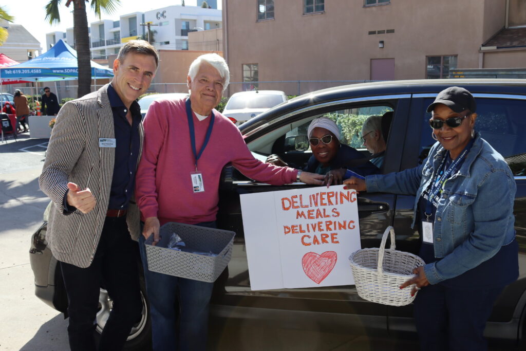CEO of Meals on Wheels San Diego County, Brent Wakefiled in a blue collared shirt and tan sports coat, standing next two a male and female volunteers holding a poster board "Delivering Meals Delivering Care" and a heart in red paint outside the driver door of a black small SUV with a female volunteering sunglasses smiling during the South County Volunteer Appreciation drive-thru event.