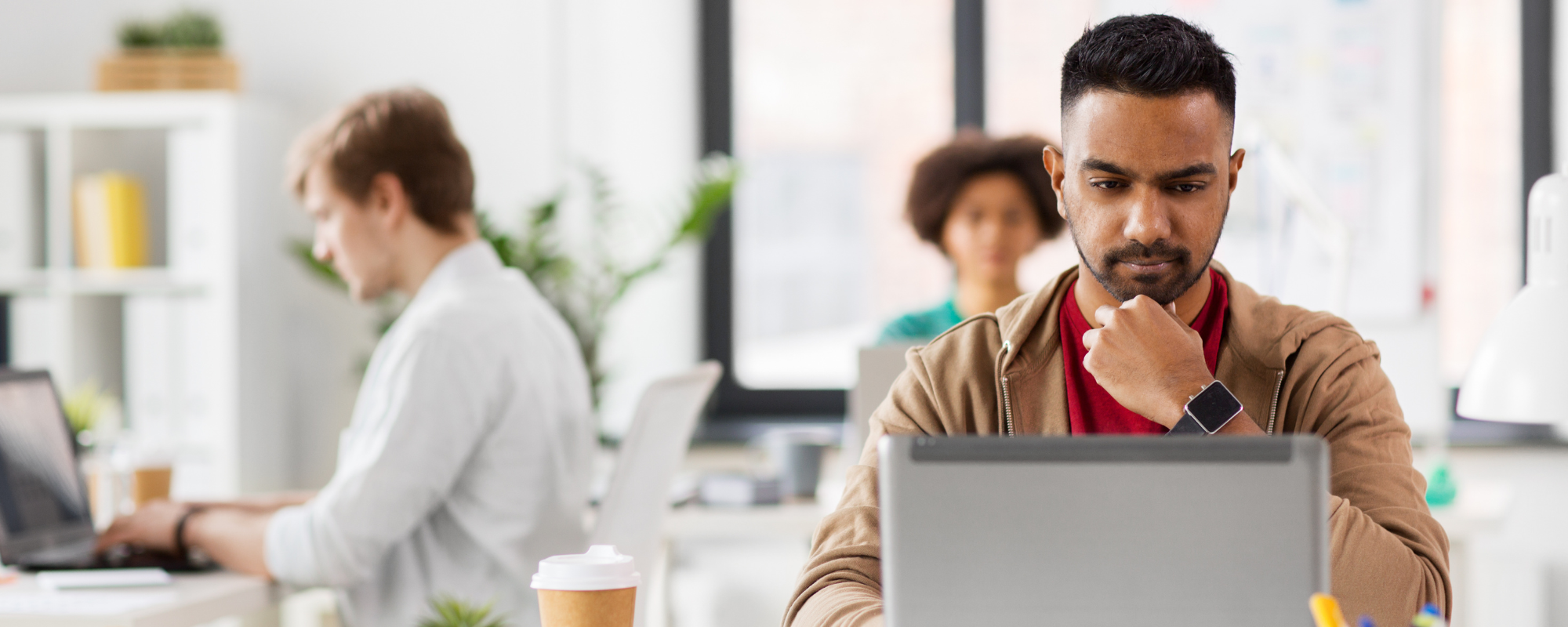 3 colleagues in an office working on computers. The focus is on the BIPOC male with black hair on the right of the image staring perplexed at his gray laptop in a red shirt, brown hoodie.