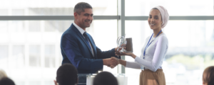 A businesswoman smiles as she receives an award from a man in a blue suit in front of her peers.