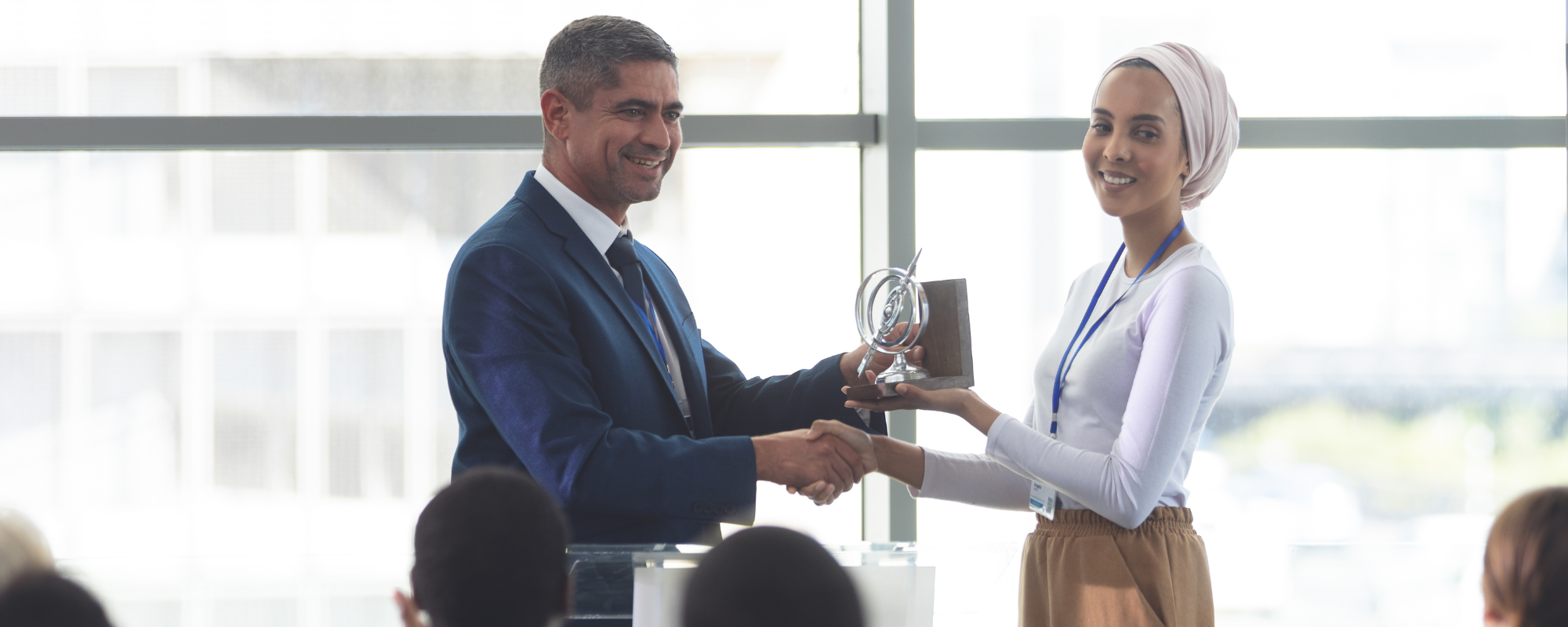 A businesswoman smiles as she receives an award from a man in a blue suit in front of her peers.