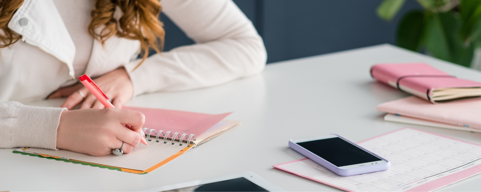 A woman holds a pink pen writing diligently on a pink notebook with a phone and tablet just within reach.